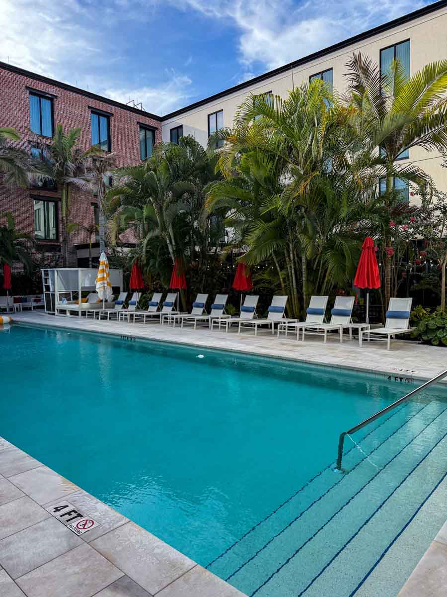 A rectangular pool with brick hotel buildings on two sides and lined with sun loungers with red parasols on a sunny day