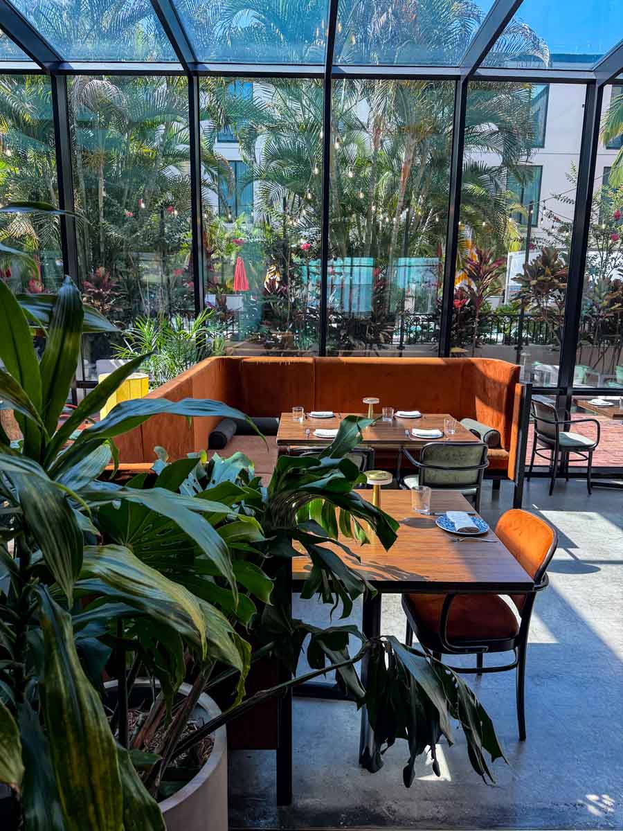 Inside a glass room at a hotel in Tampa Bay with rust orange seating and wooden tables set for eating, beyond the glass is lots of palms