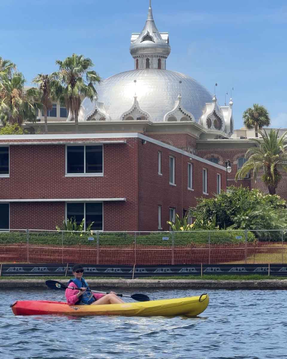 Amy in a kayak in the river with buildings and a large silver domed building behind her