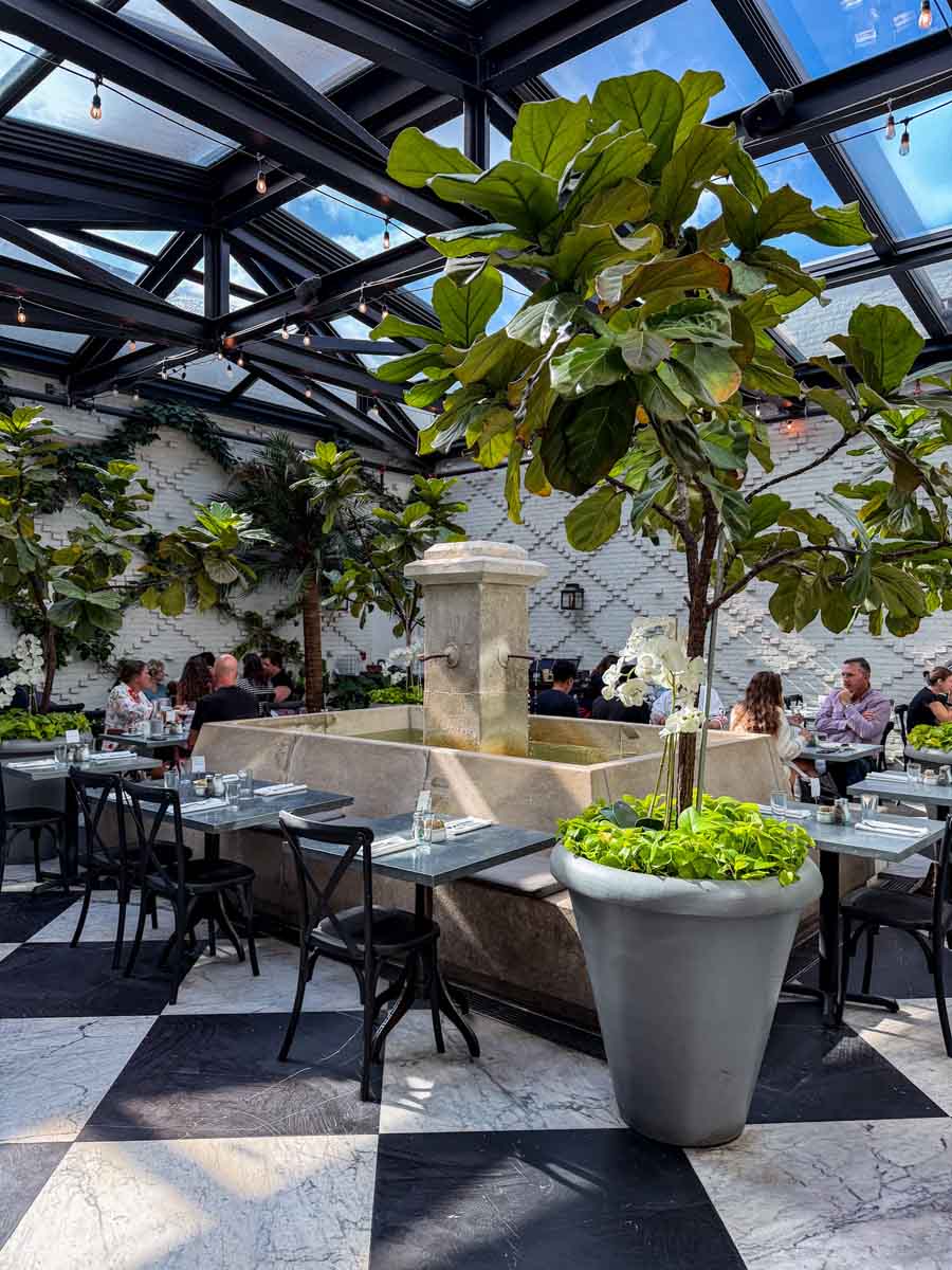 An orangery-style restaurant with glass ceilings and lots of light overlooking tables that are set up around an indoor water feature and lots of large potted trees