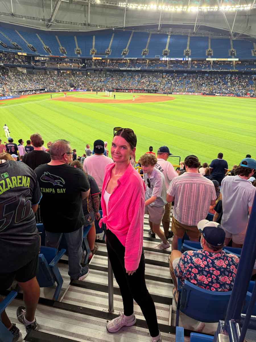 Amy standing on the stairs of a baseball stadium with crowds all around her and players on the green baseball pitch beyond her