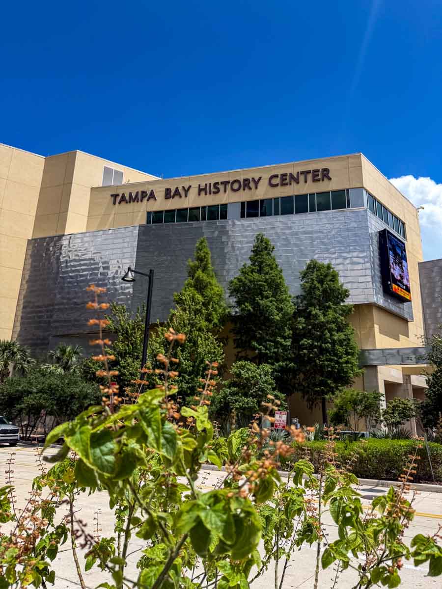 A large beige building with a portion of silver reflective surface and a sign that reads "Tampa Bay History Center"