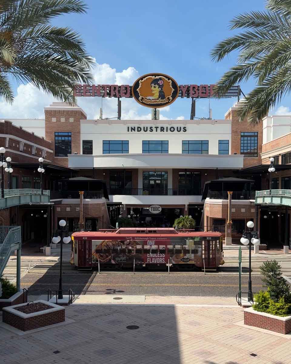 Looking down steps and across a road with a street car passing by, beyond the street car is a large shopping complex with "Centro Ybor" sign above it