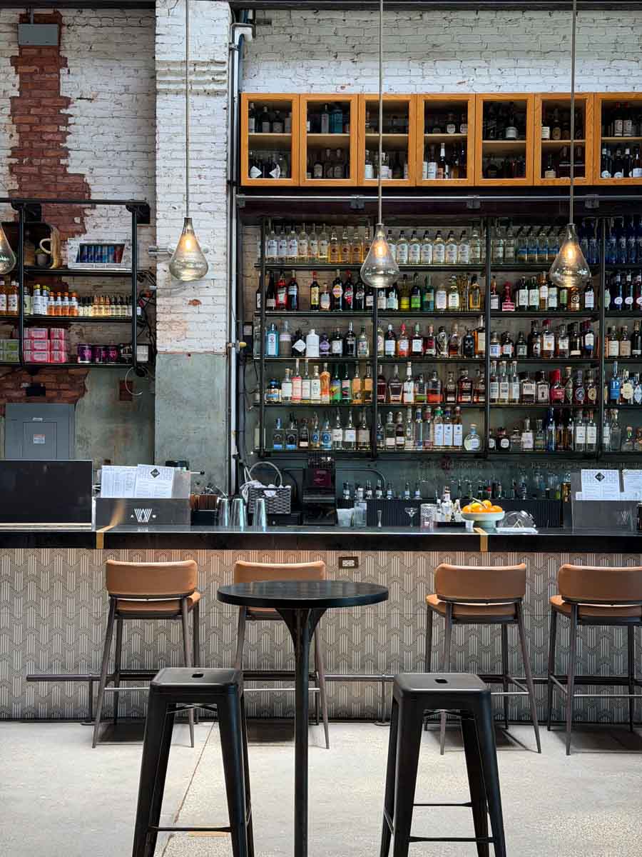 The interior of an industrial-style restaurant with 4 seats at the bar and a wall of bottles behind the bar. The wall is exposed brick