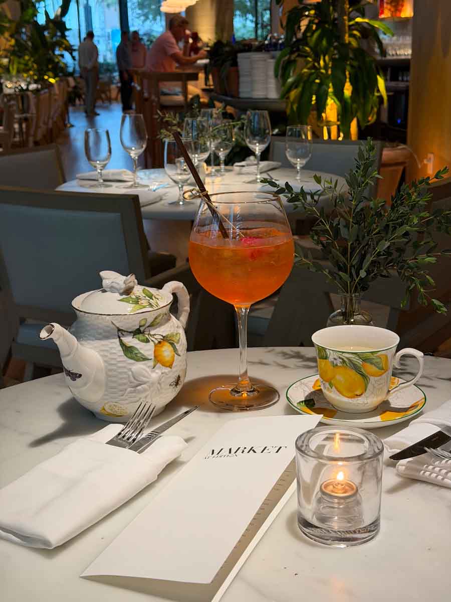 A marble table with cutlery, a cup and teapot with lemons printed on it and a large wine glass in the centre filled with orange liquid and a black straw in a restaurant