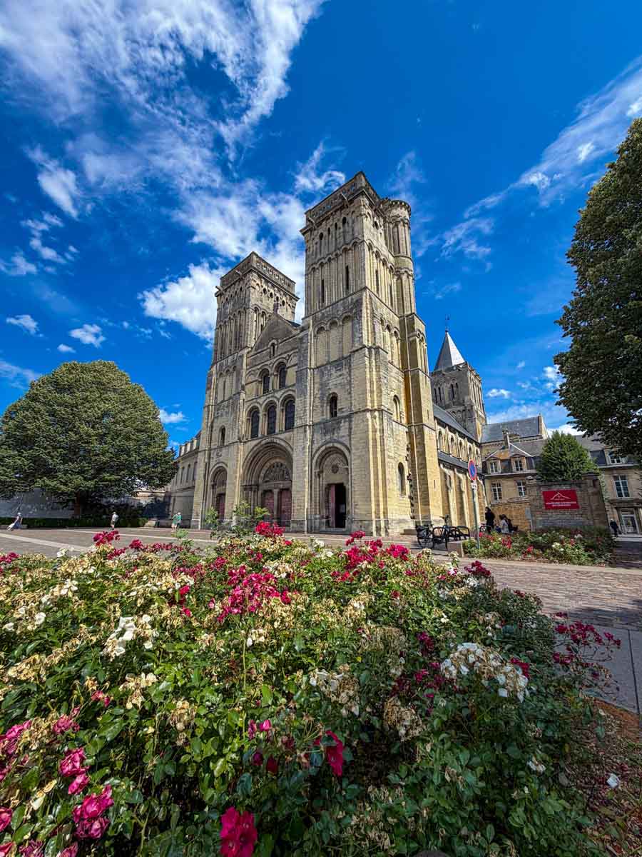 The front of a large abbey church in Caen with two towers either side of the door. There are flowers and greenery in the foreground