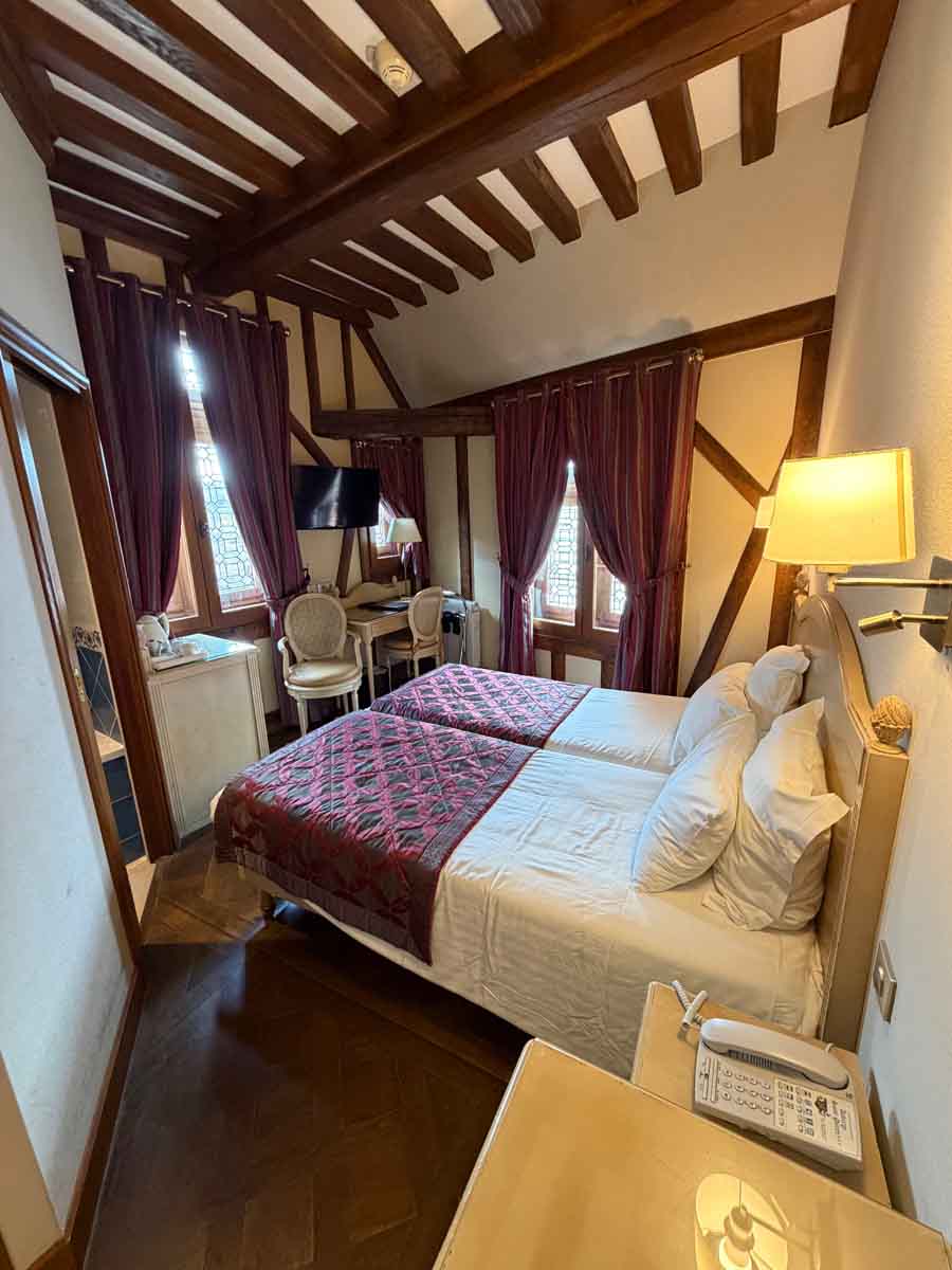 A hotel room with a double bed in the middle and two windows on the far walls. The walls and ceiling are timberframed and red curtains partially cover the windows