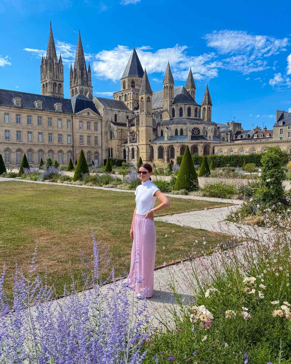 Amy standing on a path next to a mowed lawn with purple flowers in the foreground and a gothic-style castle in the background with many turrets and windows