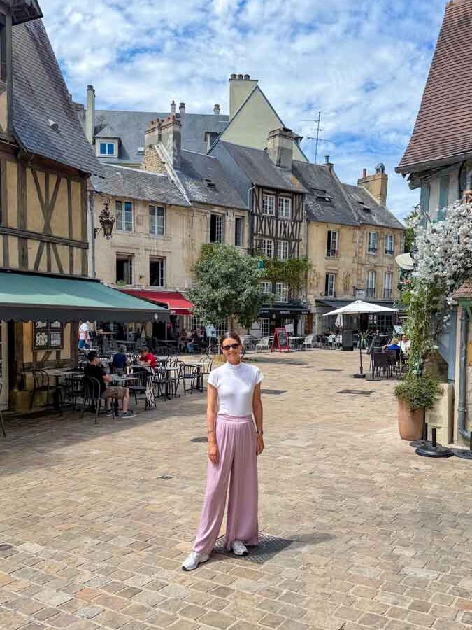 Amy standing in the middle of a cobbled square lined with old medieval style and timber-framed buildings in Caen, France
