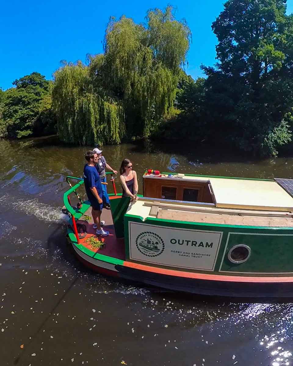 Amy and two men standing at the back of a river boat cruising along the river in Derby. It's a sunny day with green leafy trees lining the far bank