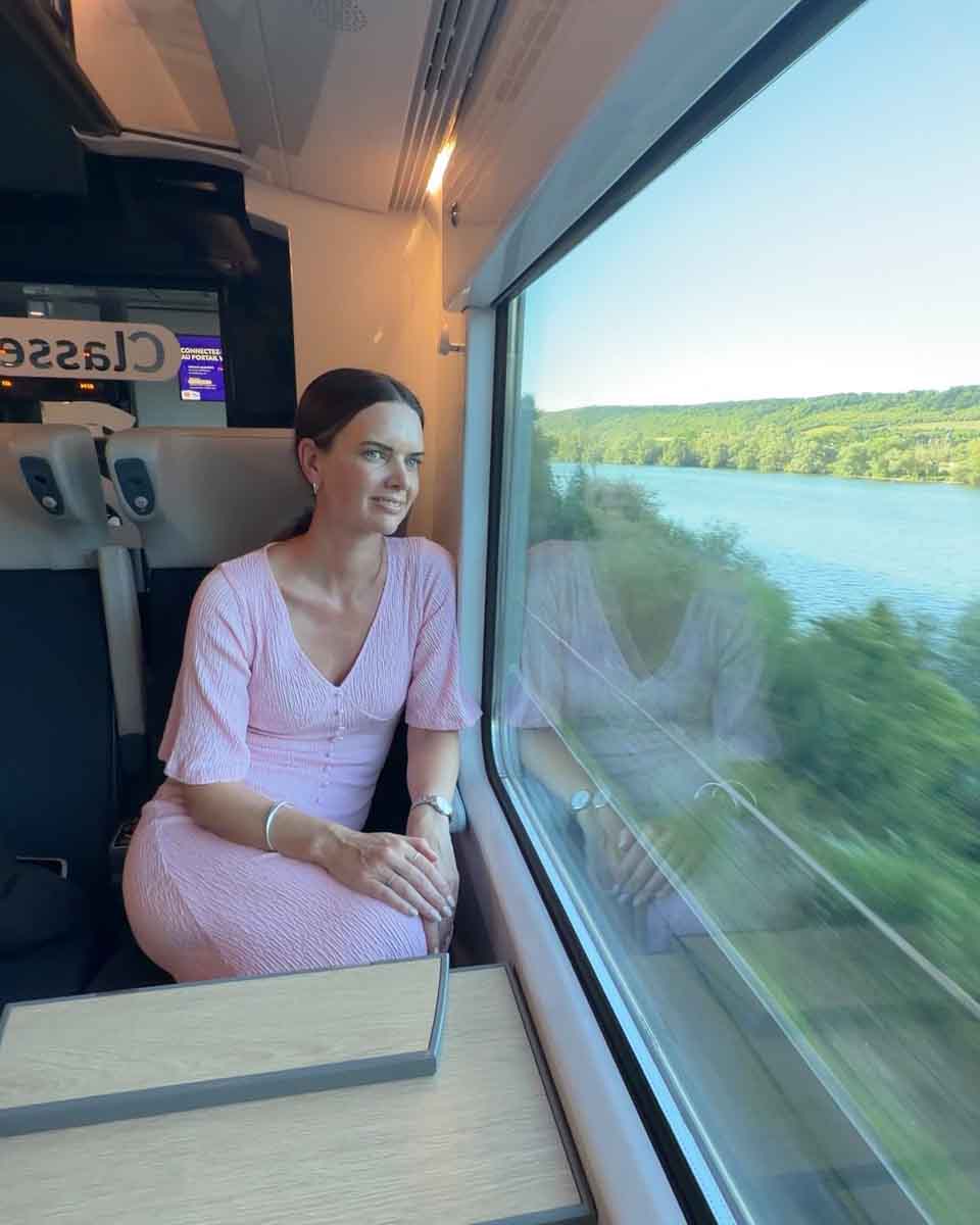 Amy in a pink dress sitting on a train seat and looking out the window at a lake and greenery going past, exploring Normandy by train from Paris