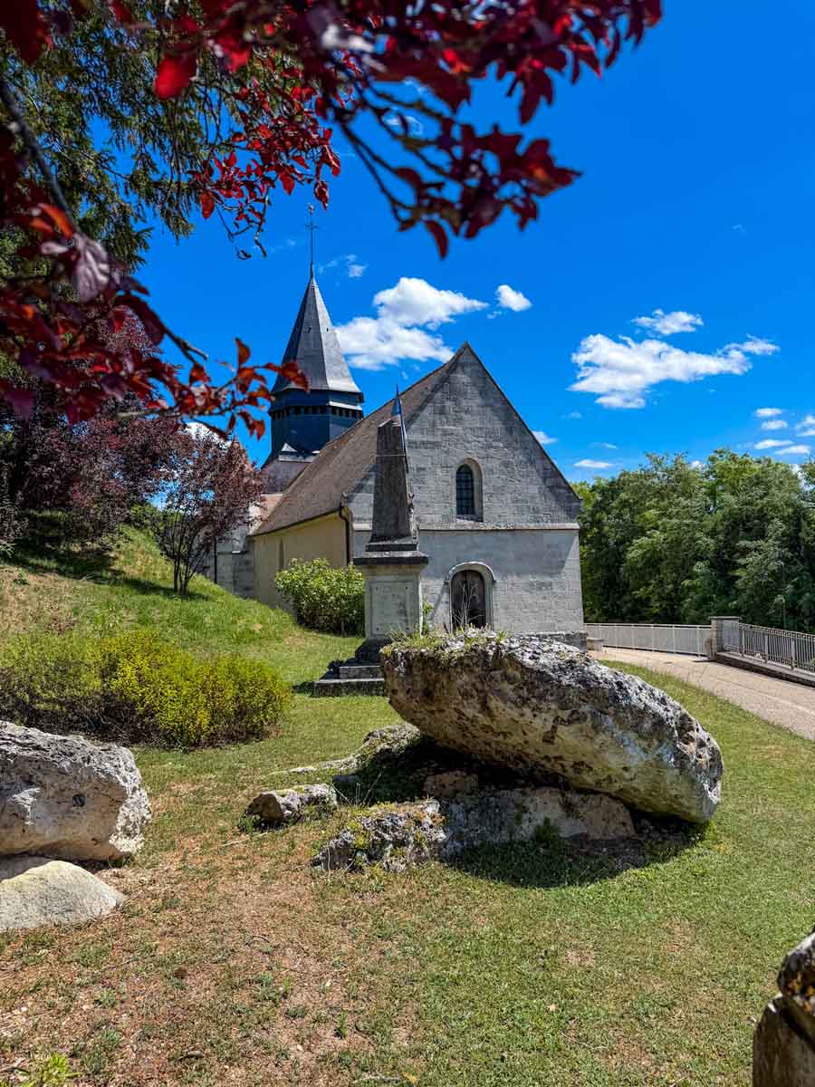A small white church with a spire and large rocks on the grassy foreground. The sky is vibrant blue with trees overhanging the left side