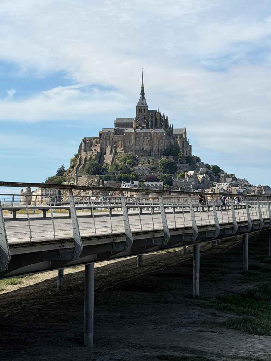 Looking across a metal walkway where beyond is the island of Mont Saint-Michel with a castle towering over the town that covers in small island