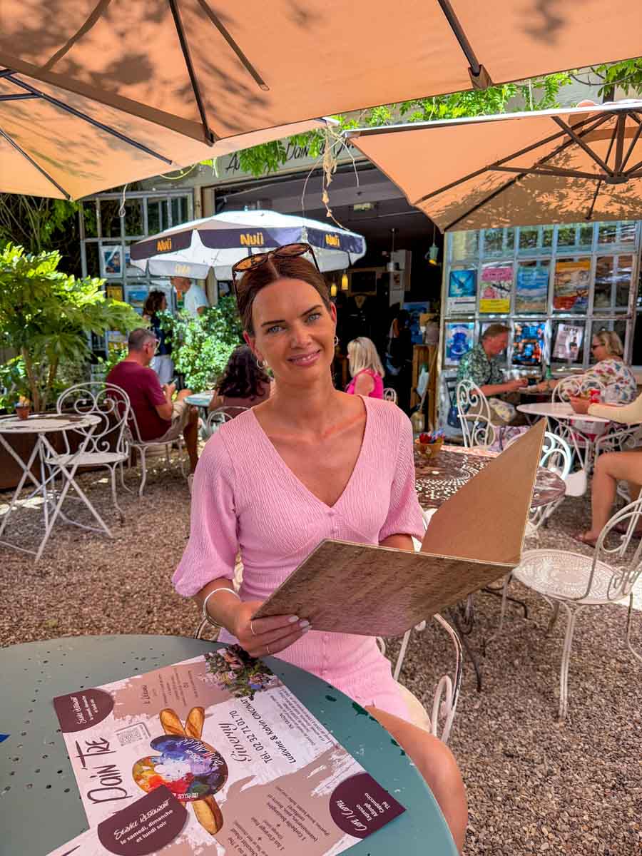 Amy in a pink dress holding a restaurant menu sitting at an outdoor restaurant table covered by a large umbrella