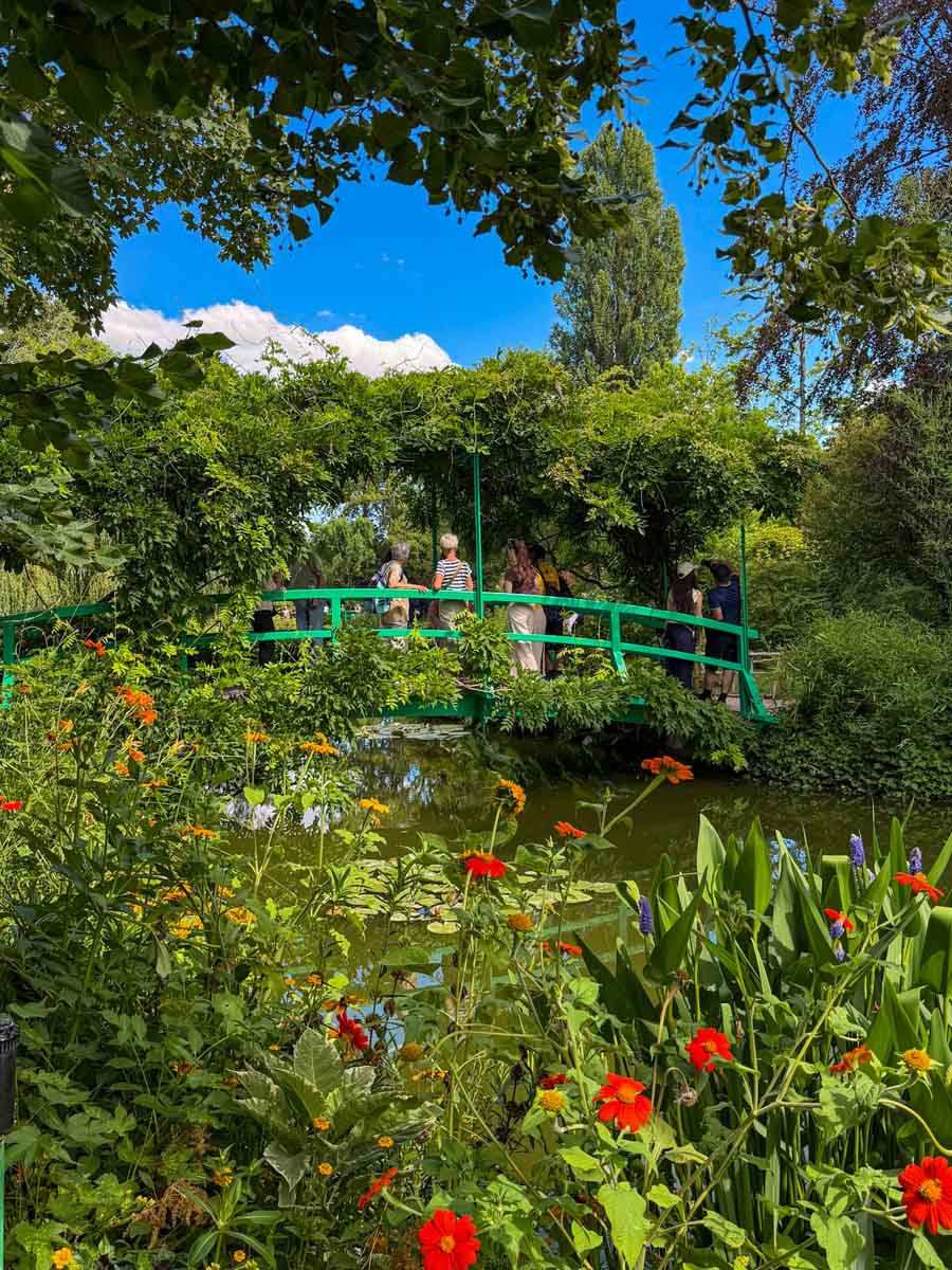 A lush green garden at Monet's House, a great day trip from Paris to Normandy on the NOMAD train. In the foreground is wildflowers and beyond is a pond with a green arached bridge over it with people standing on the bridge