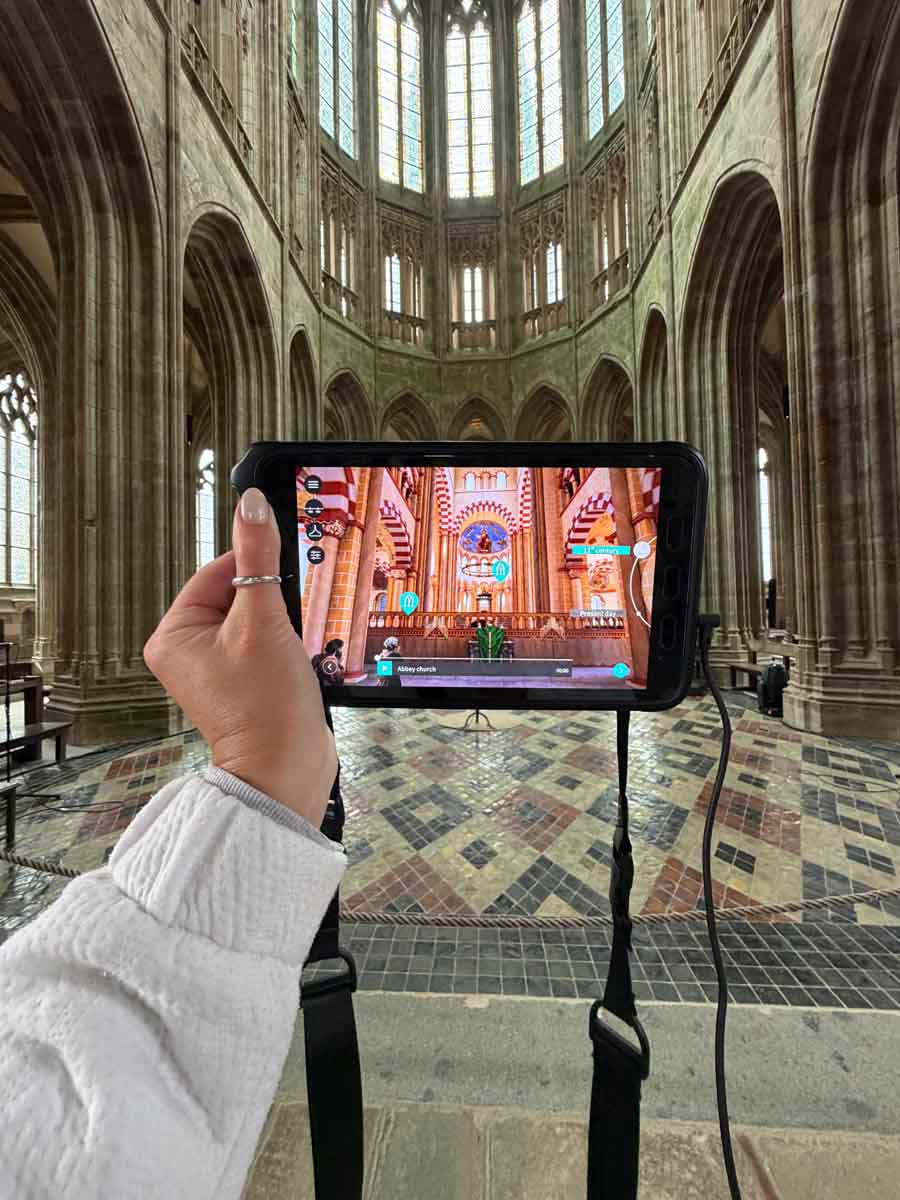A hand holding up a tablet towards the end of a church interior. The image of the tablet screen is colourful and shows an alternative4 view of the interior of the Mont Saint Michel Abbey