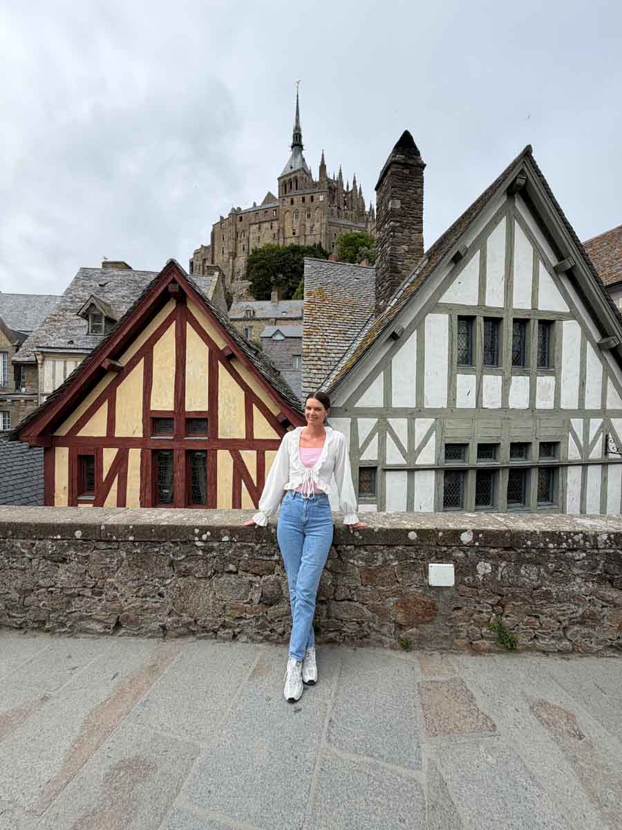Amy leaning against a wall with a view of timber-framed houses behind her and the turrets of a castle in the distance