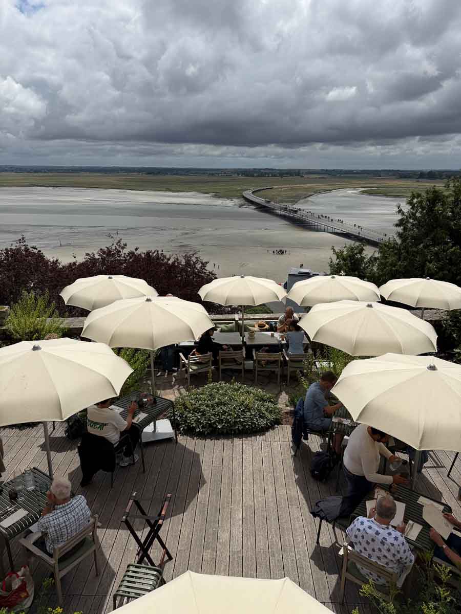 Lookingdown on a wooden deck of a restaurant covered in parasols with poeple dining beneath them. Beyond is a view of a coastline with the tide out on a cloudy day