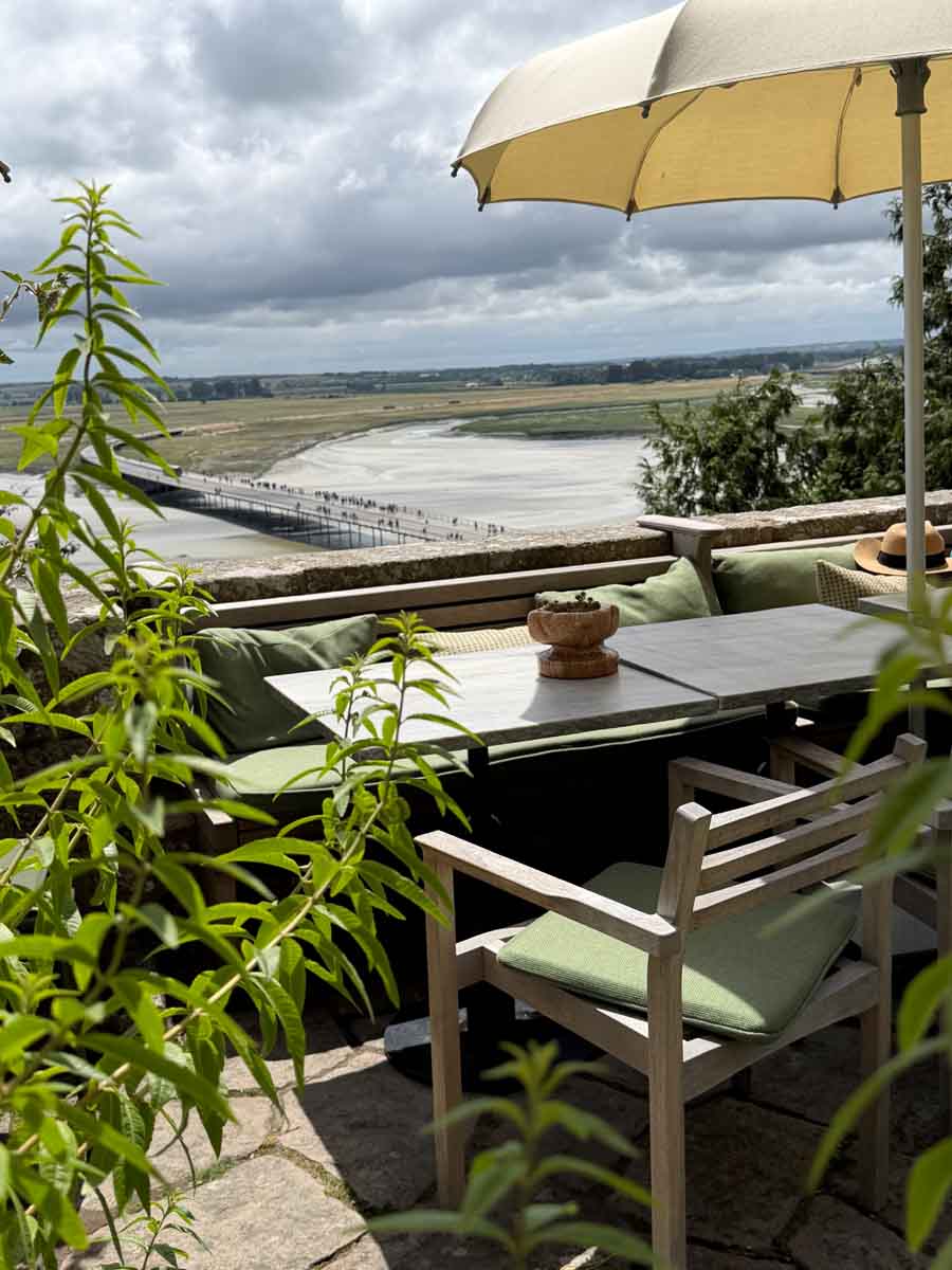 A restaurant in Mont Saint-Michel outside on a terrace overlooking the bay. The table is surrounded by seating with grrn cushions an a parasol overhead
