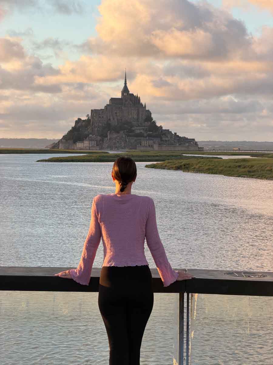 Amy standing on a viewpoint looking out across a bay of water with the dramatic island of Mont Saint-Michel beyond. The sky is partially cloudy and the clouds are orange as if it's sunset