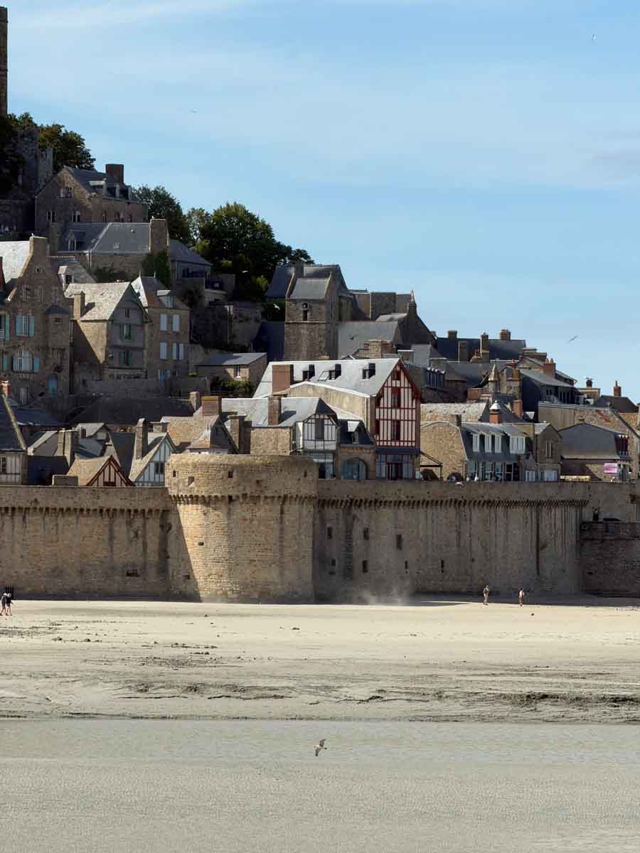Looking across a sandy beach to where stone fortress walls tower over the sand and a medieval town with many stone and timber-frame buildings sit on the hillside above the fortress walls of MontSaint-Michel in France