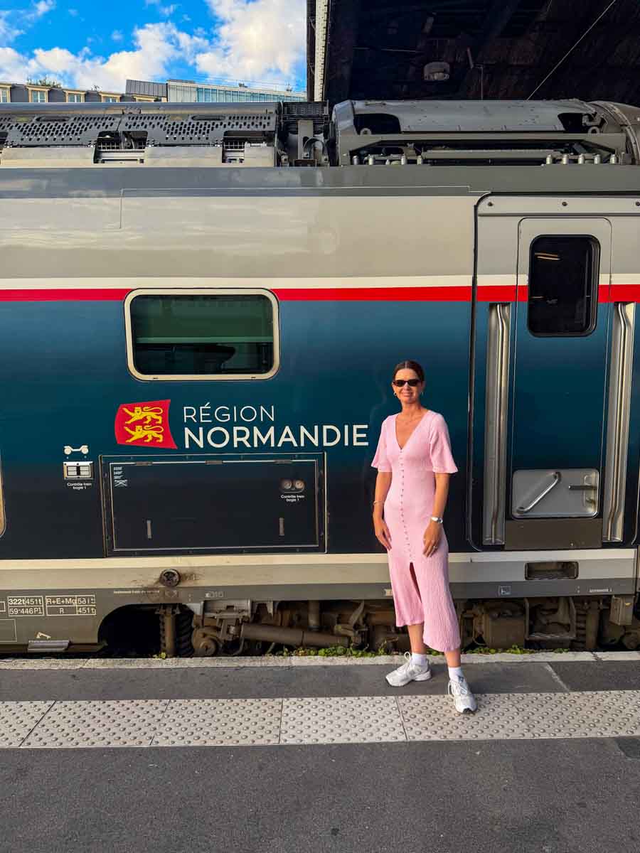 Amy in a pink dress standing in front of a train that is stationary on the platform. The side of the train reads "region Normandie" - the NOMAD train to visit Normandy by train from Paris