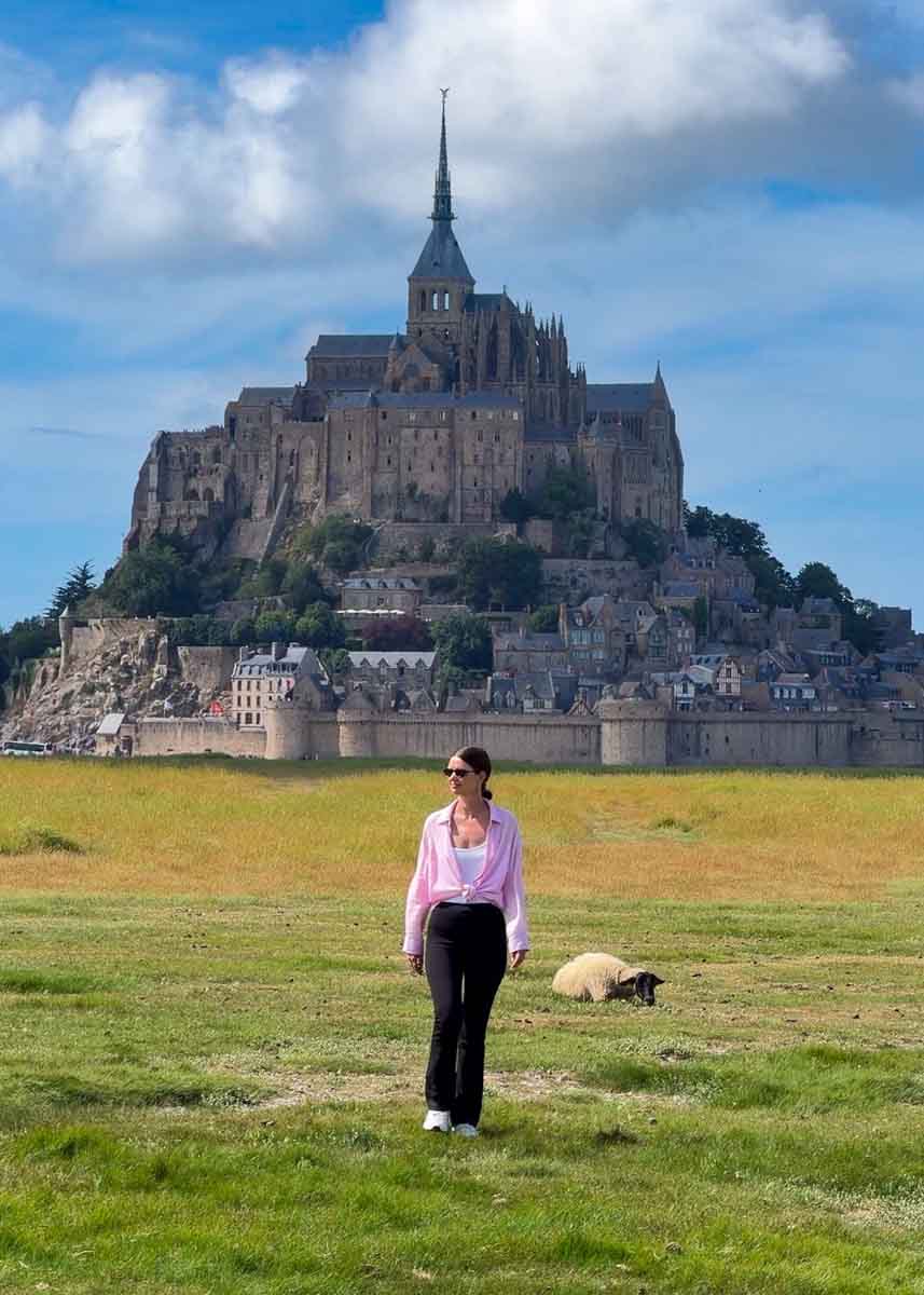 Amy walking across grass with a single sheet behind her and beyond that is the towering island of Mont Saint-Michel, surrounded by fortress walls and covered in stone and timber-frame houses with a dramatic castle perched on the top of the island