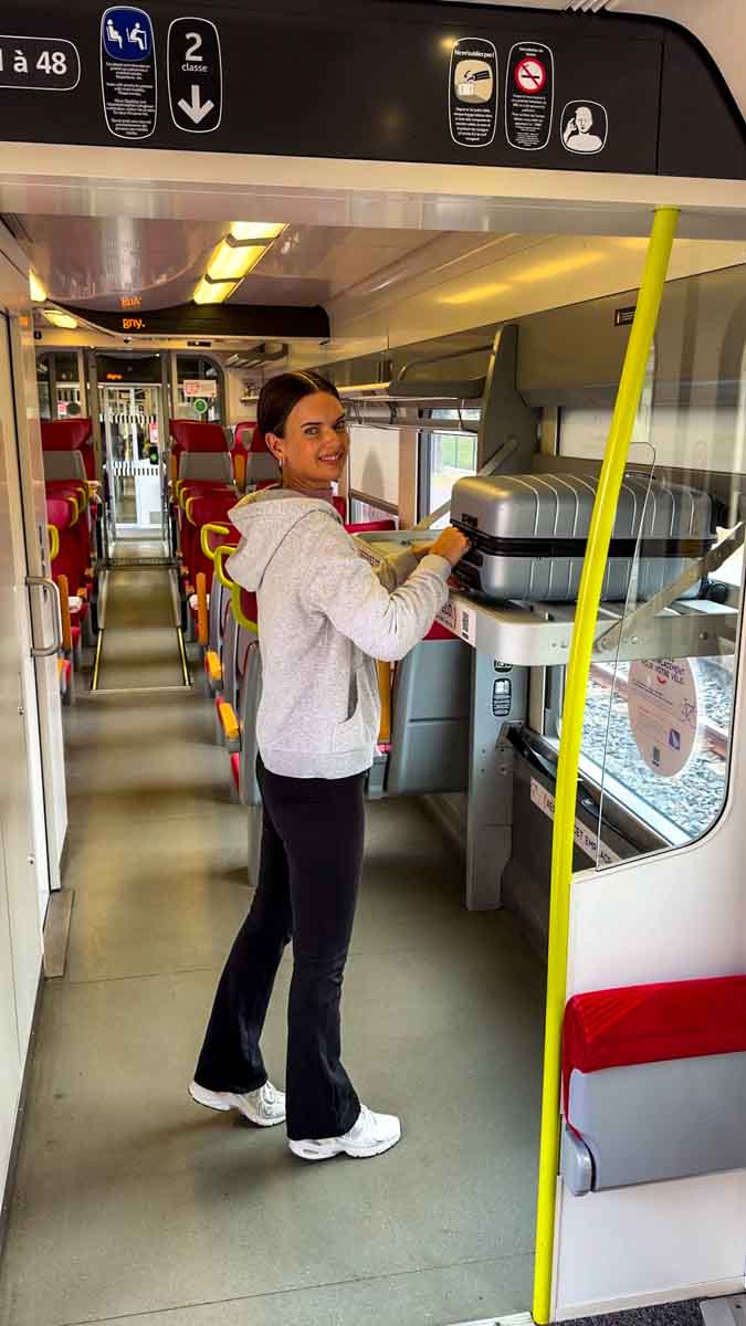 Amy on a train, loading her silver suitcase onto the luggage rack and smiling at the camera while visiting Normandy by train fom Paris